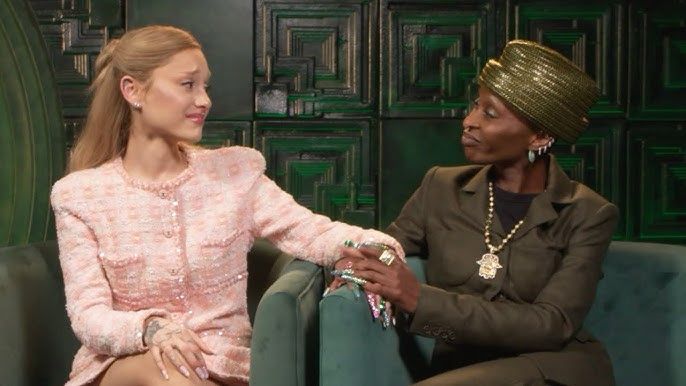 Ariana Grande in a pink suit seated on a green chair and Cynthia Ervio in a dark green suit seated on a green chair beside her. They are holding hands and looking at one another.