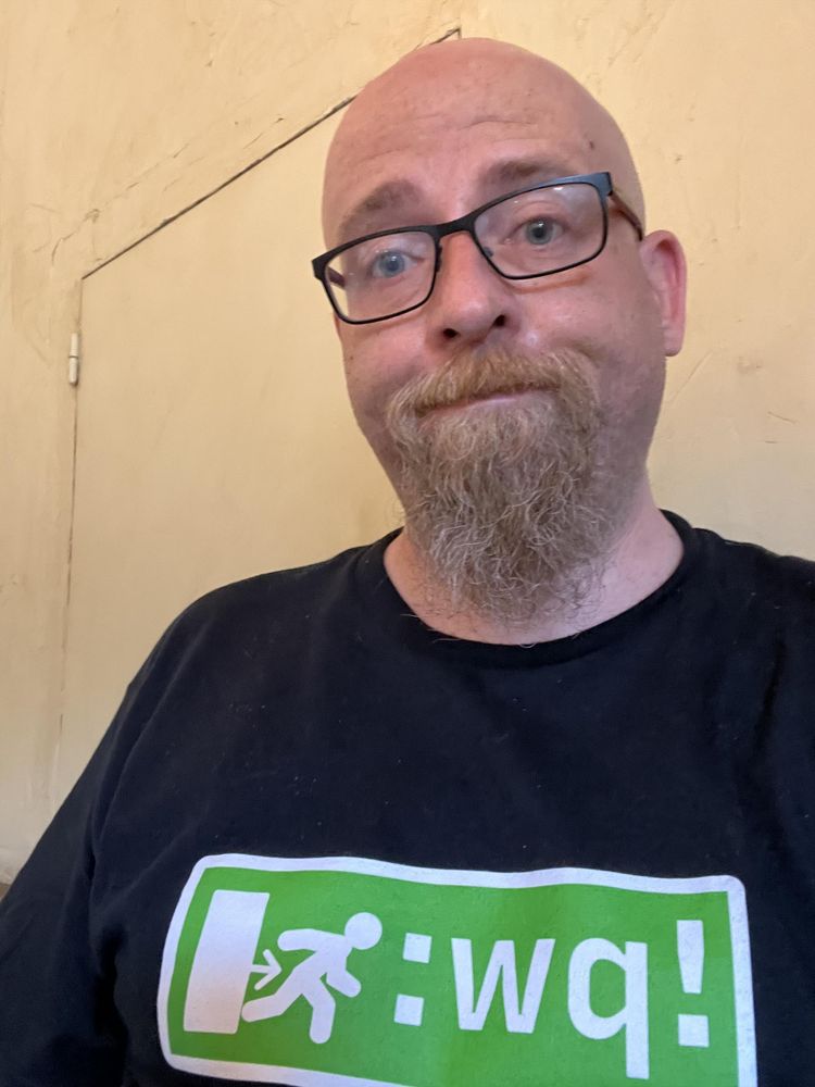 Moi, chauve barbu a lunettes, portant un t-shirt noir avec le signe « sortie » qu’il y a au dessus des issues de secours, avec un pictogramme et texte en blanc sur fond vert. Sauf que le texte est :wq! , une des commandes pour sortir de vim