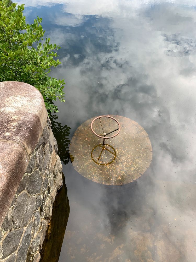 Photo of a valve in concrete maybe, surrounded by water, with a stone and cement wall to the left. The sky and clouds are reflected in the water, making it look almost like smoke. The whole thing looks rather surreal.
