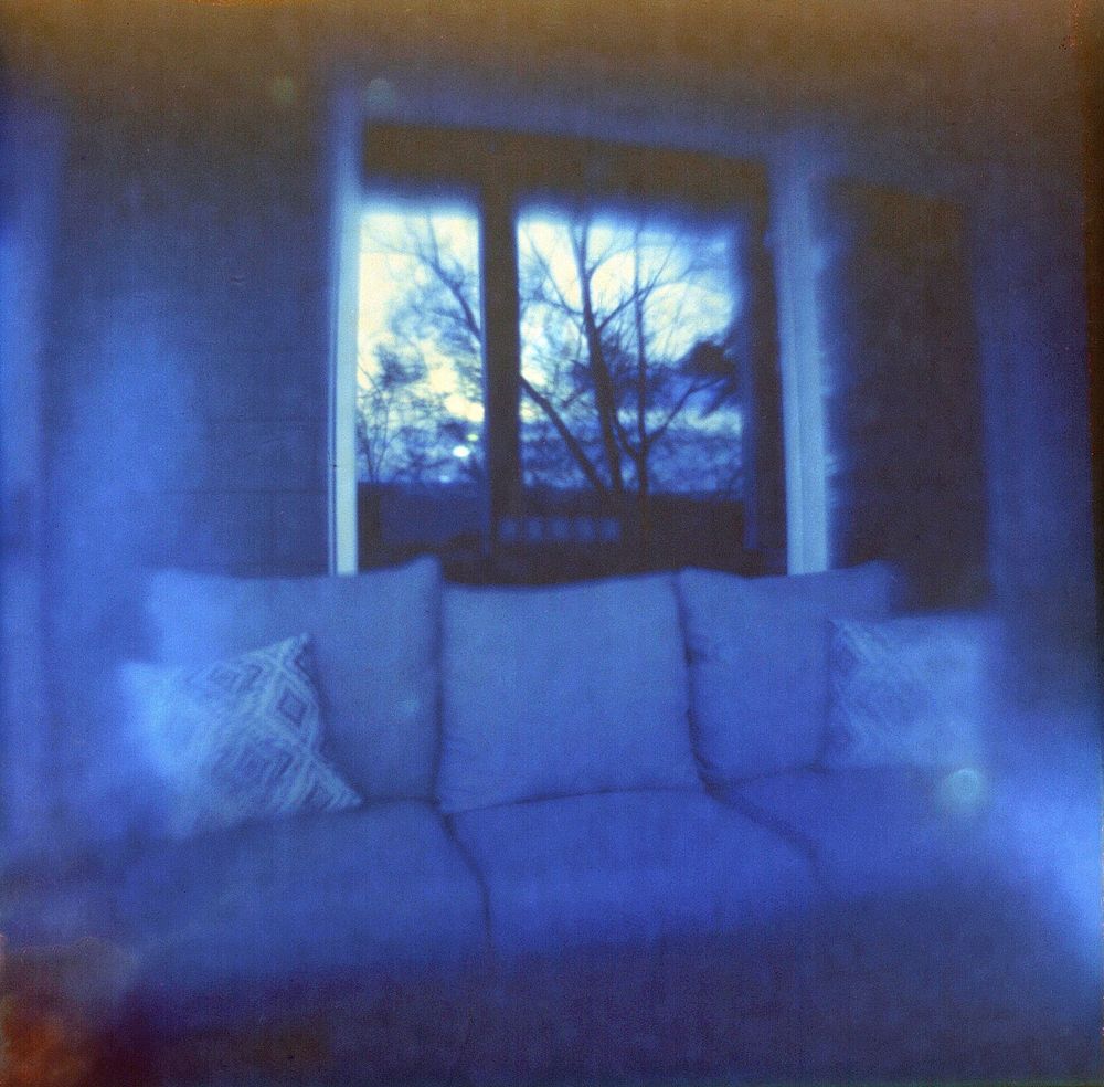 Photo of a couch with blurry edges, trees are reflected in the large glass window behind the couch. The whole photo has a bluish tint.