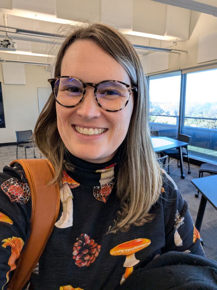 A woman in a mushroom-patterned top and glasses is standing in a university classroom, smiling