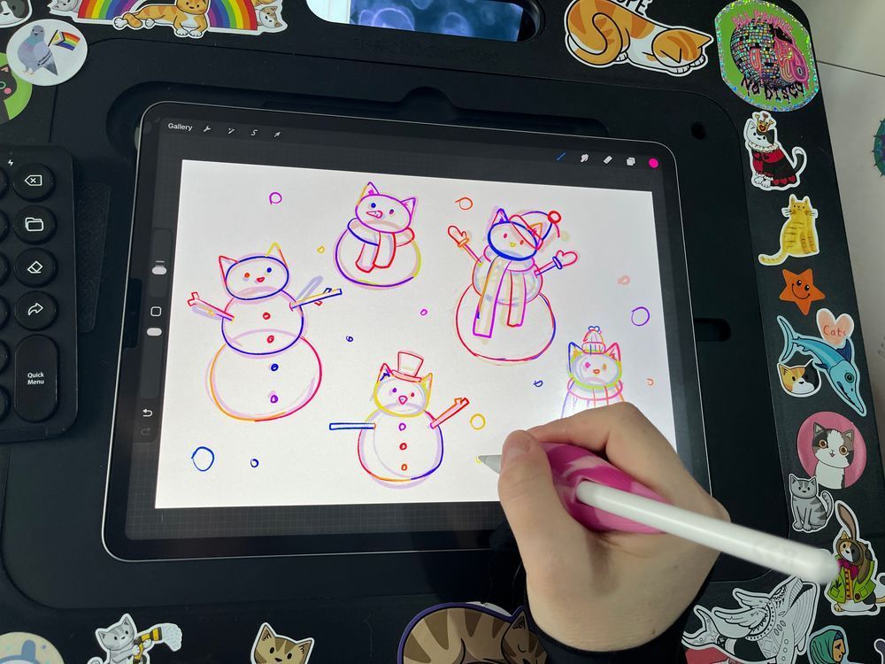 a hand holding a pencil over an ipad, drawing snowcats in procreate. the iPad is mounted in a large board covered in stickers