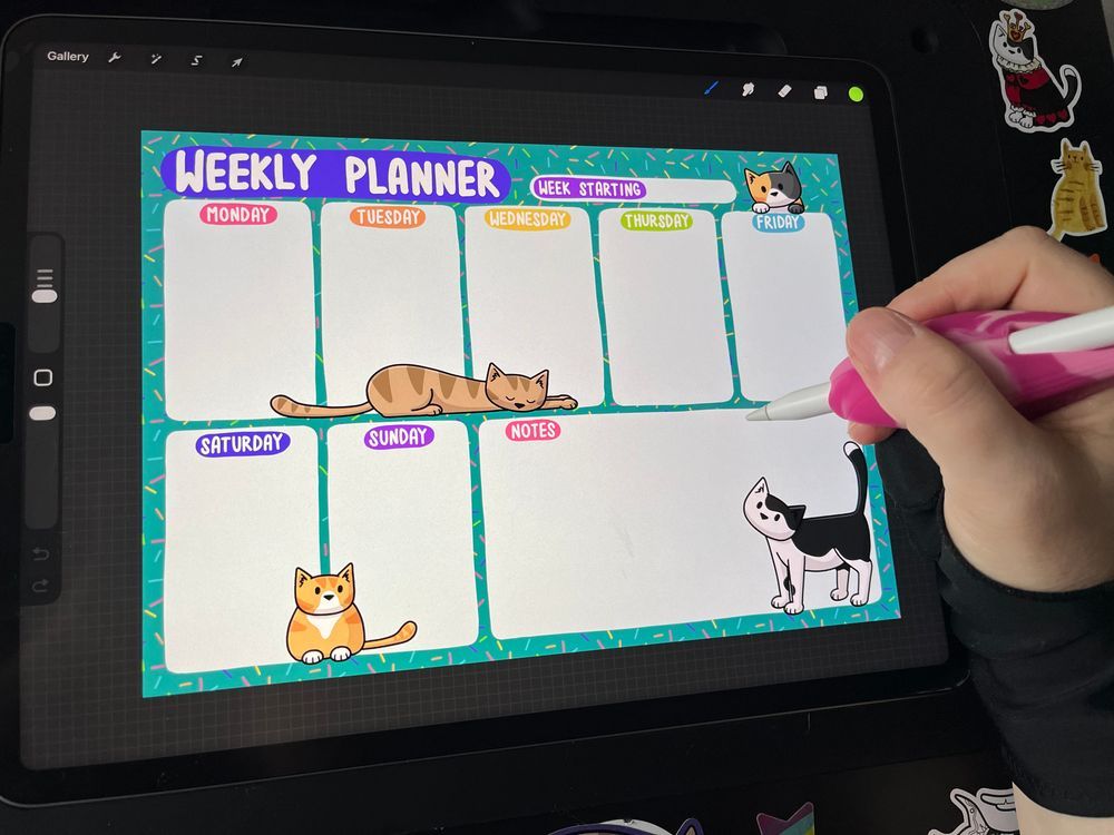 a hand drawing on an iPad. there's a colourful weekly planner on the screen, adorned with cartoon cats