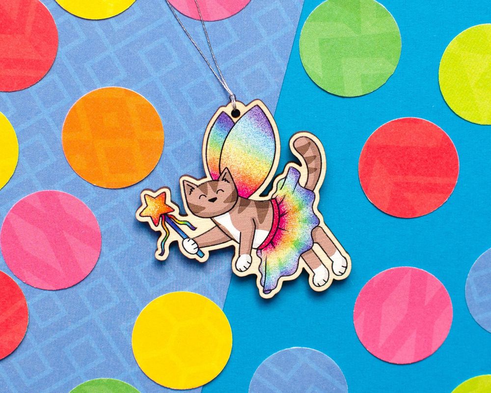 a wooden decoration - a cartoon cat dressed in rainbow tutu and wings, with a sparkly star wand, printed on wood with a silver string to hang it. it's on a blue paper background with colourful circles