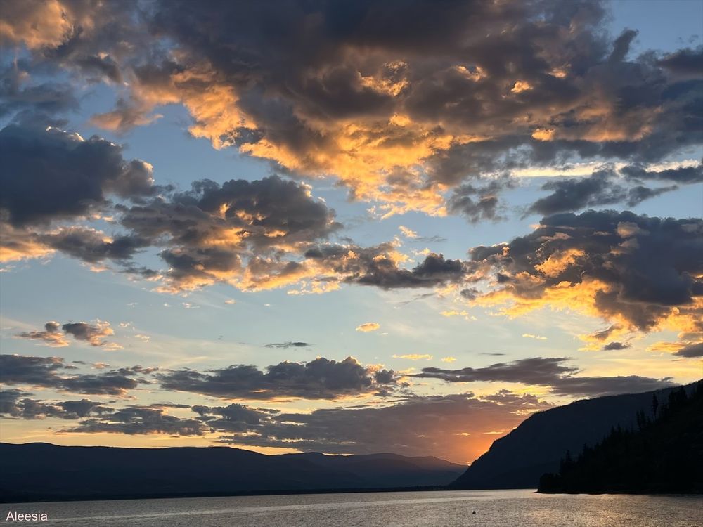 The sun coming up over Shuswap Lake in Chase, British Columbia. The sky is blue and orange with scattered clouds. There are several dark mountains in the distance.