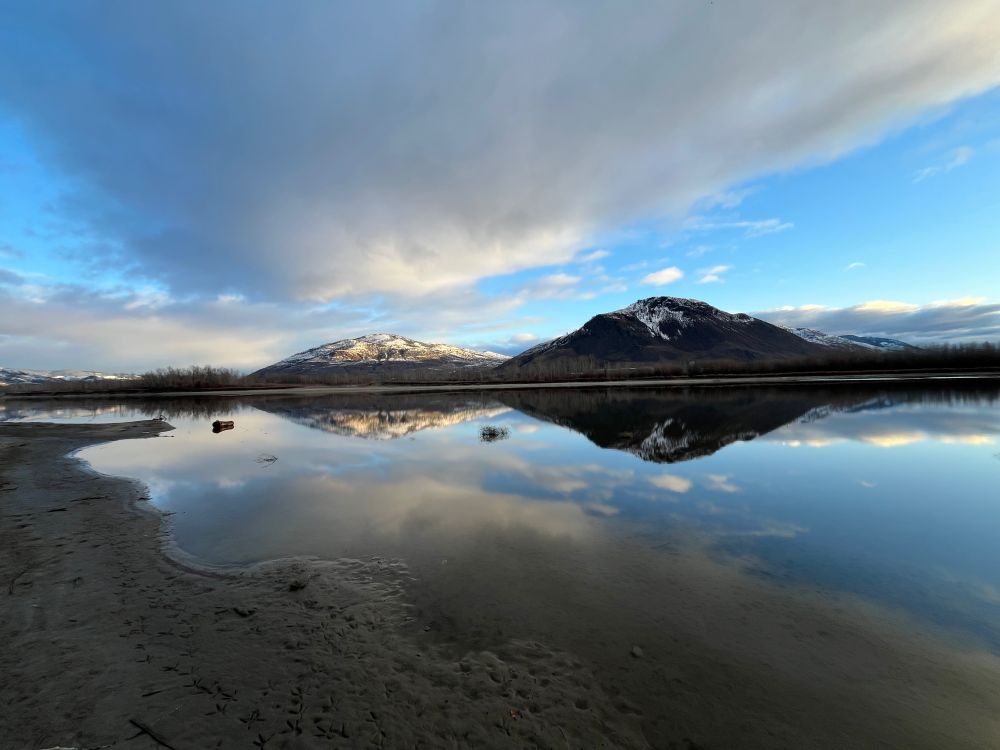 A large cloud hovering over the Thompson River in the Interior of British Columbia. The sky is blue and there are snow capped mountains in the distance. There’s a mirror image on the water beside the sandy beach.