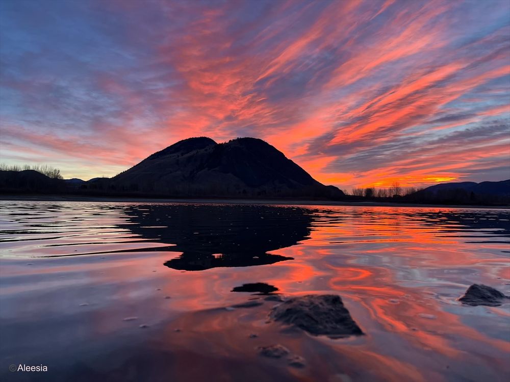 The sun coming up over the Thompson River, in the interior of British Columbia. The sky is beautiful shades of blue, pink and orange. There are dark mountains in the distance, and a few small rocks near the shore. There’s a mirror image on the water.