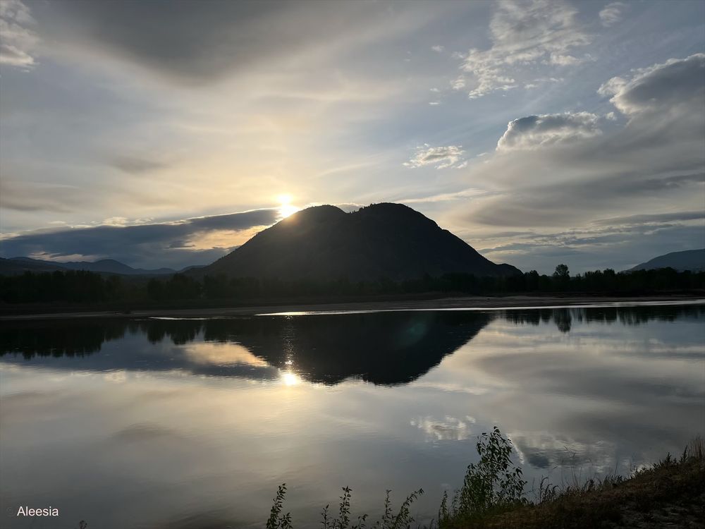 The sun coming up over the Thompson River, in the interior of British Columbia. There are scattered clouds, but the sky is starting to clear. The mountains and trees are dark, and there’s a mirror reflection on the water.