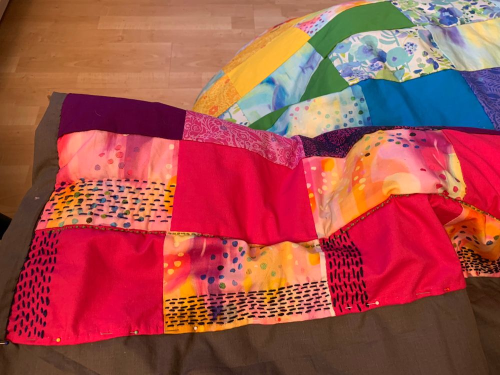 Squares of fabric in shades of pink, blue, green and yellow with lines of embroidery stitching , pinned to grey fabric background. 