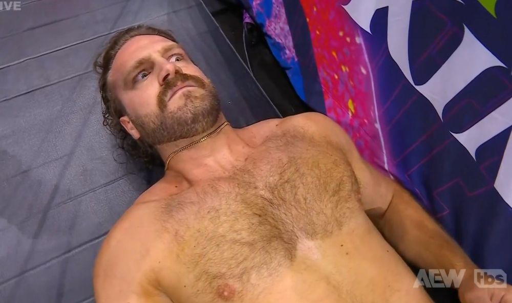 Hangman Adam Page, shirtless, lies next to a wrestling ring with a bewildered look in his eyes