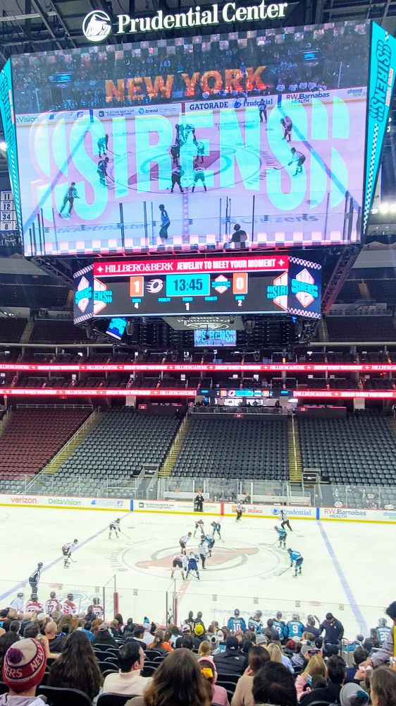 Photo taken from the stands of a hockey game between the New York Sirens and the Ottaw Charge of the PWHL as the teams take a faceoff at center ice.