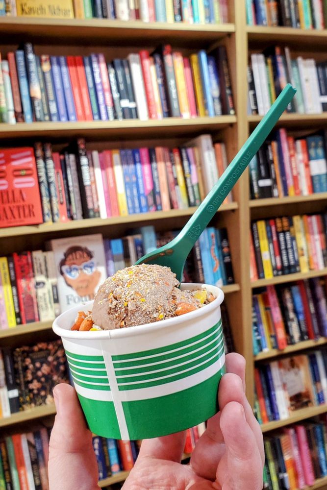 A hand holding a small cup of chocolate ice cream with Reese's Pieces topping up in front of a bookshelf filled with colorful books