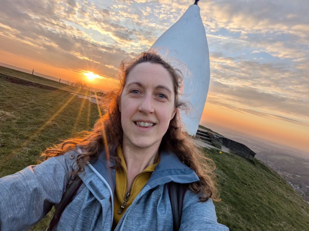 Sunset on the top of a hill with a tall white structure (known as White Nancy) and a smiling Lucy