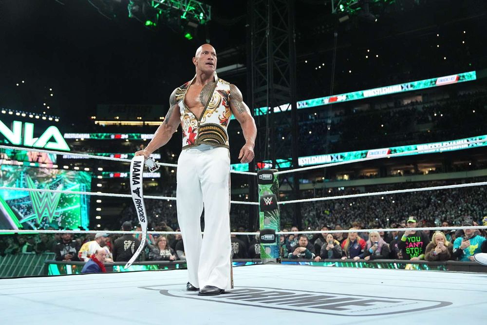 The Rock, a muscular man in a white suit, in the middle of a ring. He's wielding a white belt that says "Mama Rhodes. 