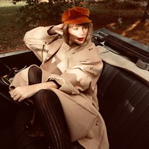 Taylor Swift sitting in a car with opened roof, wearing a beige coating and a red cap-like hat looking to the side.