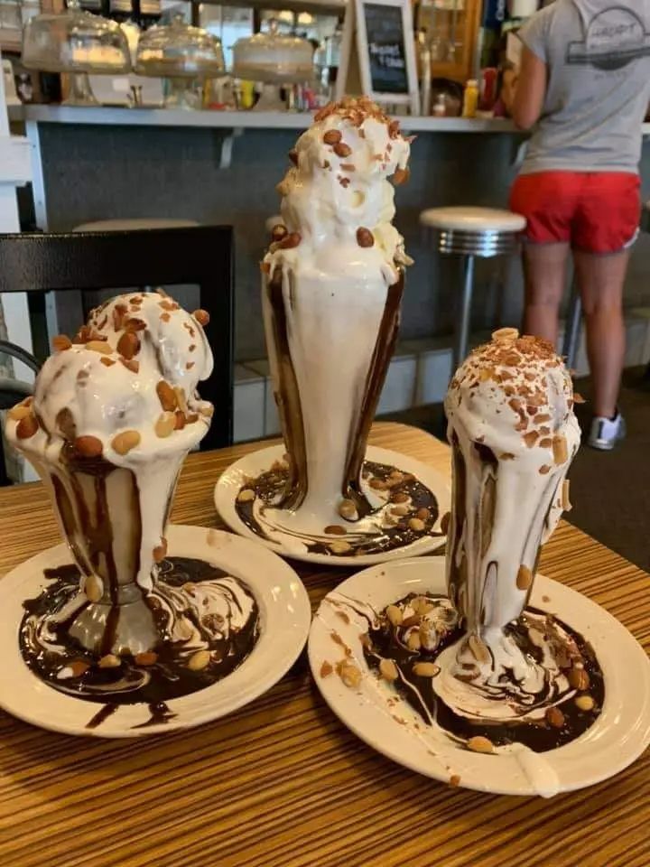 Three ice cream sundaes that are topped with chocolate fudge and melted marshmallow to the point that it's pouring all the way down the glasses and into a saucer below them