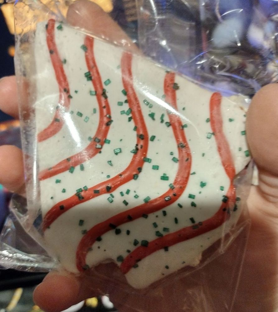 A christmas tree shaped cake wrapped in plastic. It's white frosting coated with red stripes and green sprinkles and look almost twice the size of the original ones.