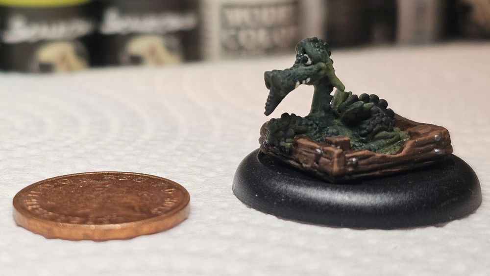 A photograph of the small green flared nostril swamp dragon Errol from the Discworld book Guards! Guards!. He is sitting in a brown wooden crate.

There is a penny to the side, which is approximately the same width across as the miniature.