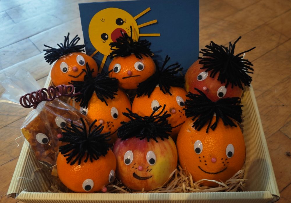 Ein kleiner Korb mit Äpfeln, Orangen und Mandarinen, die alle mit Gesichtern und Haaren aus Wolle versehen sind und die Betrachter*innen anschauen. 