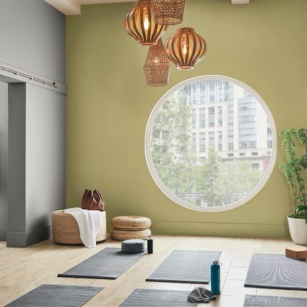 Picture of a light, green-yellow wall with a large, round window. Floor has 6 gray yoga mats, a wicker table and stool and a large plant