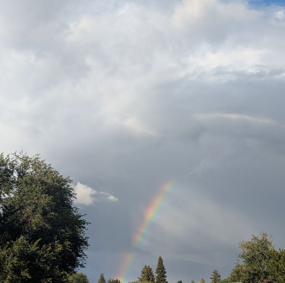a photo of a rainbow set against a grey, cloudy background. there are rays of light breaking through the clouds that appear to be shining down from the rainbow.