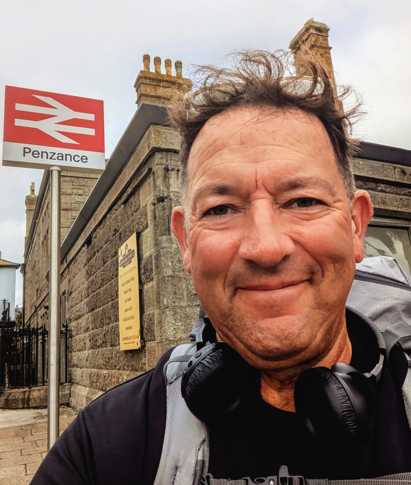 Middle aged man in black t-shirt, with a big rucksack stood in front of sign outside Penzance rail station 