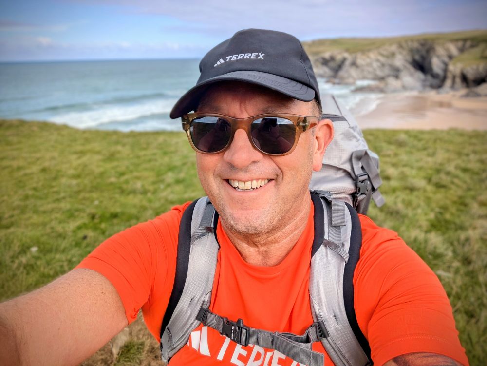 Middle aged man in orange t-shirt, cap, sunglasses with a big rucksack on a clifftop with a beach behind 