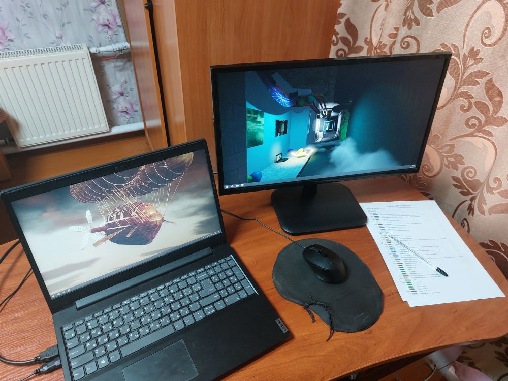 A photo of a desk with Lenovo laptop (showing screenshot from Revision 2025 invitation demo), monitor (showing screenshot from Revision 2024 invitation demo - will be replaced with Paintover), mouse on a pad and printed schedule (with black pen)