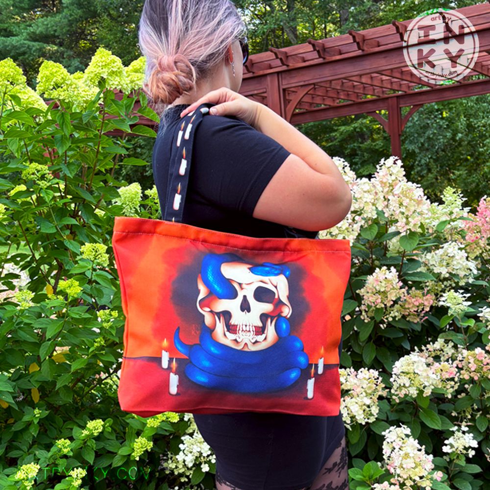 A person standing facing away from the camera with the blue snake and skull tote bag over their shoulder. They are in a black outfit against a bush of flowers.