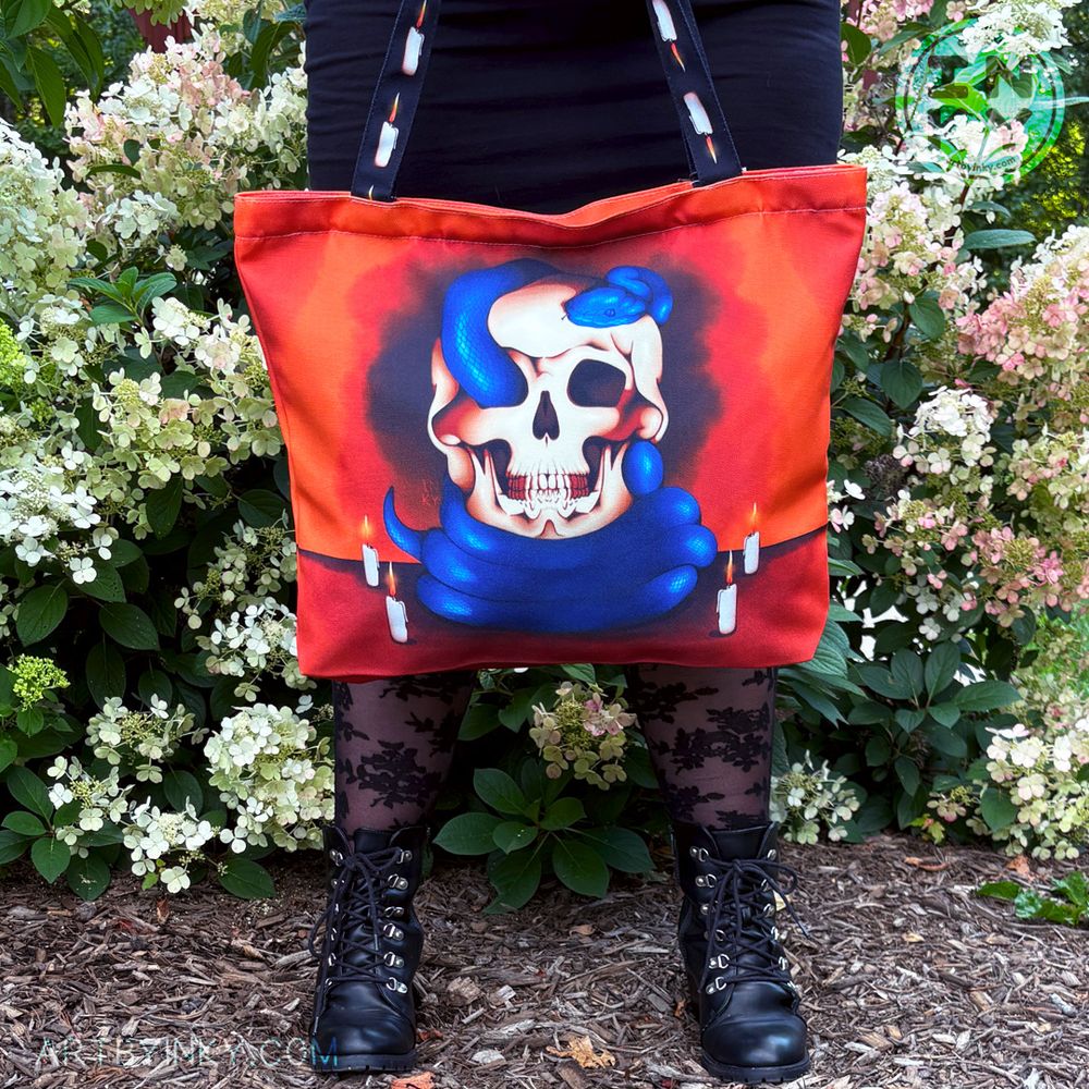 A person holding the blue snake and skull tote bag above their black boots. They are in a black outfit against a bush of flowers.