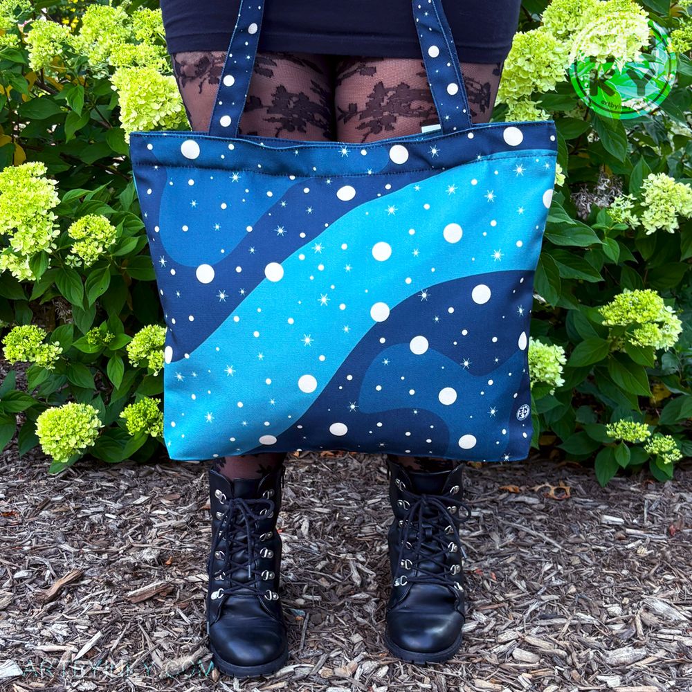A person holding the blue galaxy tote bag above their black boots. They are in a black outfit against a bush of flowers.