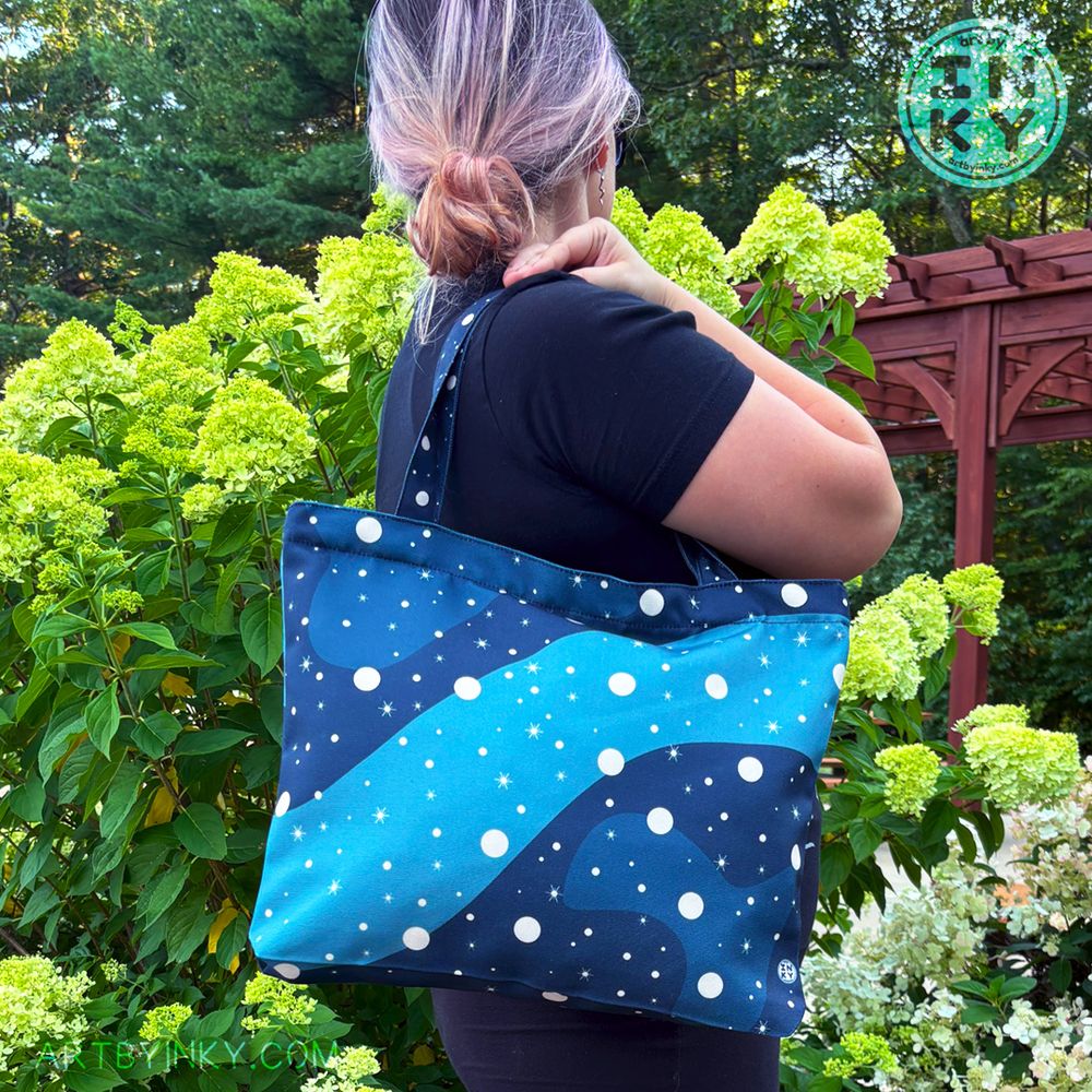 A person standing facing away from the camera with the blue galaxy tote bag over their shoulder. They are in a black outfit against a bush of flowers.