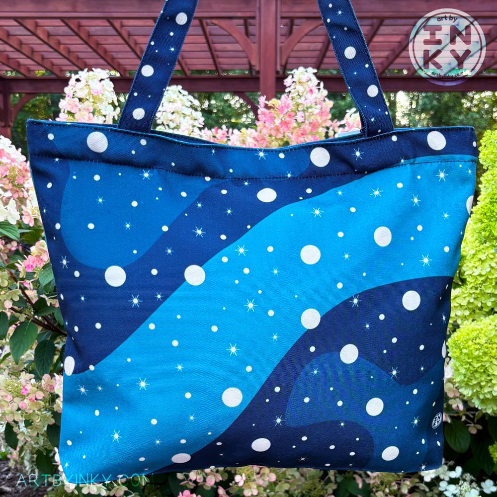 A blue galaxy tote bag against a bush of flowers.