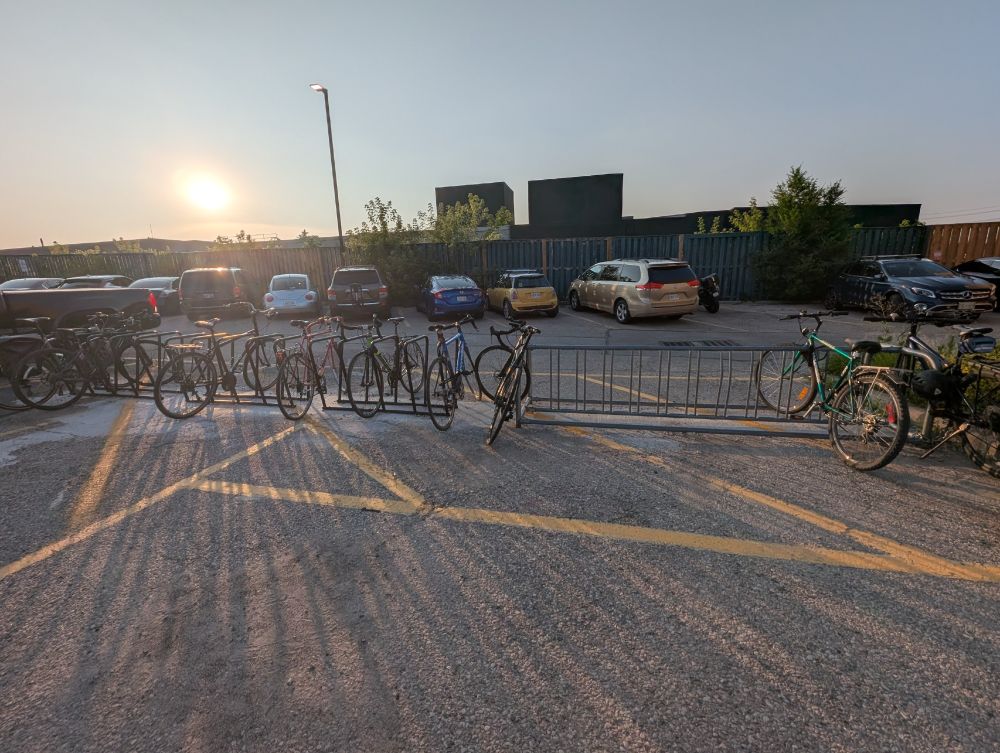 A sun-drenched parking lot at either dawn or dusk, with a long metal bike rack in the foreground. The section of the rack featuring an inverted-U design is crowded with numerous bicycles, while the simpler wave-style section to the right has only three bikes. Parked cars and a modern building are visible in the background against a clear sky.