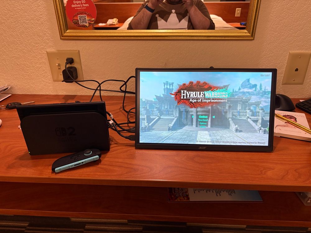 Picture of a Switch 2 on a desk next to a 16-inch portable display. The title screen for Hyrule Warriors: Age of Imprisonment is on the display