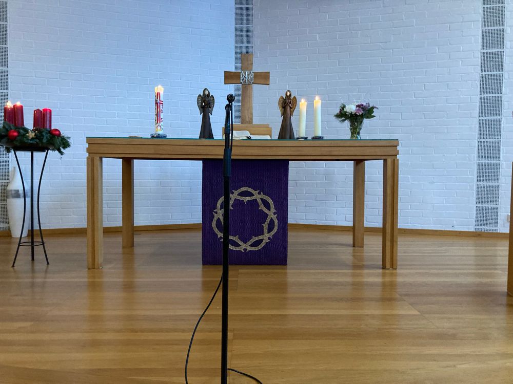 Altarraum Erlöserkirche: Lila Antependium für Advent. Kreuz, Bibel und Kerzen auf dem Altar, außerdem Blumenschmuck. Neben Kreuz und Bibel stehen rechts und links Engel. Neben dem Altar ein Adventskranz mit drei brennenden Kerzen 