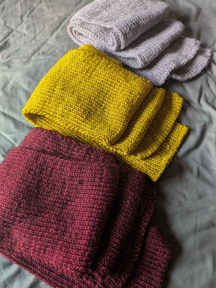 3 hand-knit scarves laid out vertically. in order from top-to-bottom, the colors are lavender, yellow, and dark red.