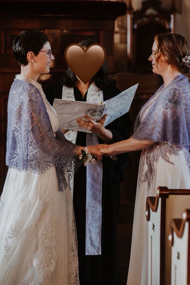 Two women in lace shawls and wedding dresses holding hands during their ceremony