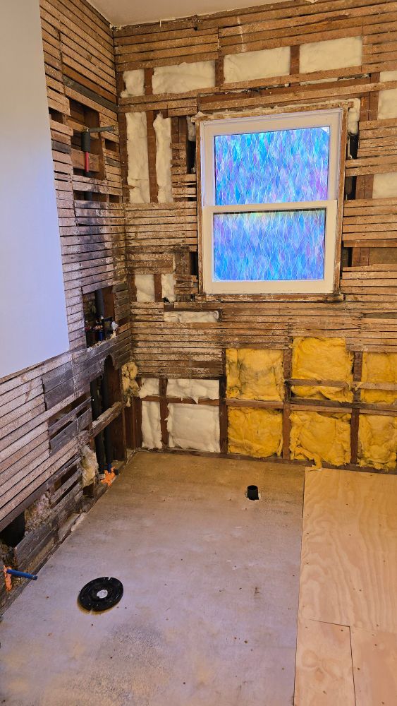 Room with a window and exposed lath, a pipe for the shower drain and toilet ring on the floor, and insulation in the walls, which makes a change from the nothing that was in there before lol