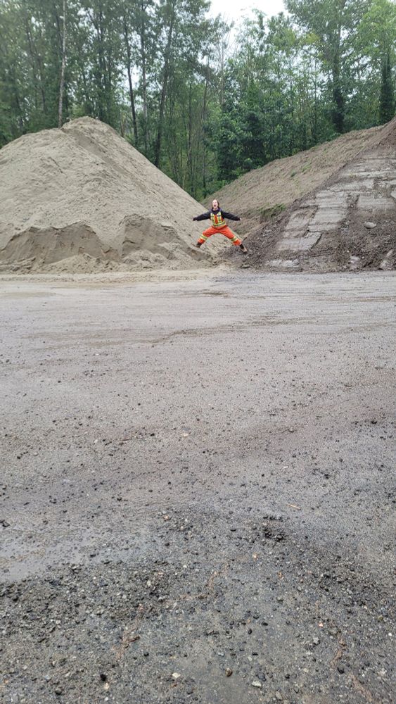 Photo of a person in the distance in orange coveralls and a black hoodie, standing right between two massive piles of sand and imitating the "I am the sand guardian" Vine. 