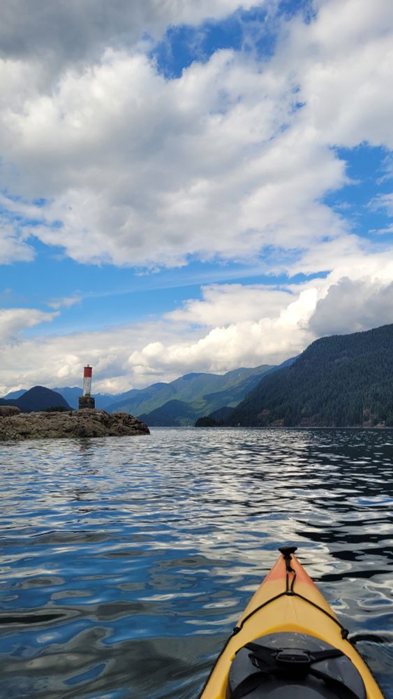 Photo of an ocean bay with mountains in the background. The sky is a mix of sun and cloud. There is a small island with a tiny lighthouse on the left side. The nose of a yellow kayak is visible in the bottom half of the image. 