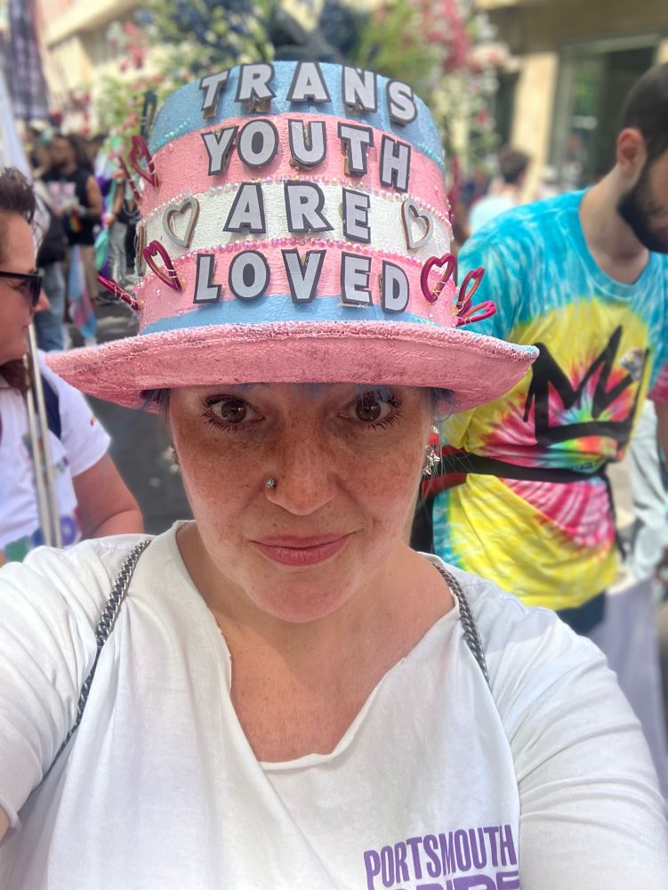 Abi at Pride in London 2024, wearing a Portsmouth Pride logo tee and a top hat painted like a trans flag and decorated with thy flowers and hearts and the slogan TRANS YOUTH ARE LOVED