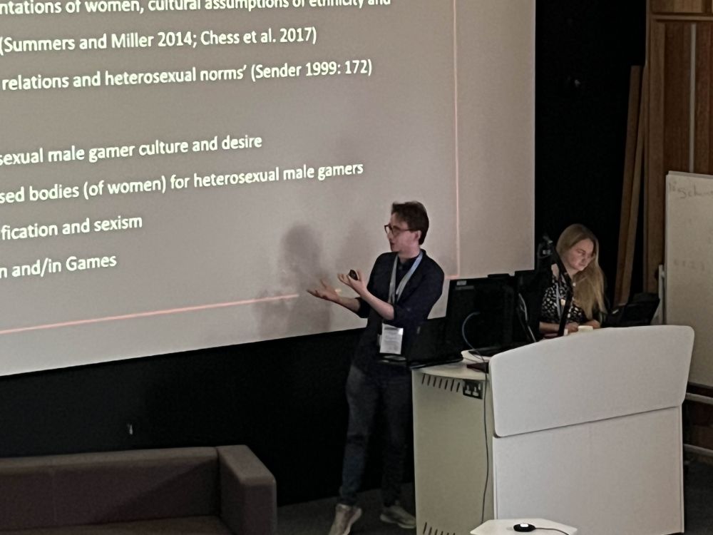 Man in black shirt and woman with blonde hair behind a white podium, with words on the screen behind them including the phrase 'heterosexual male gamers'.