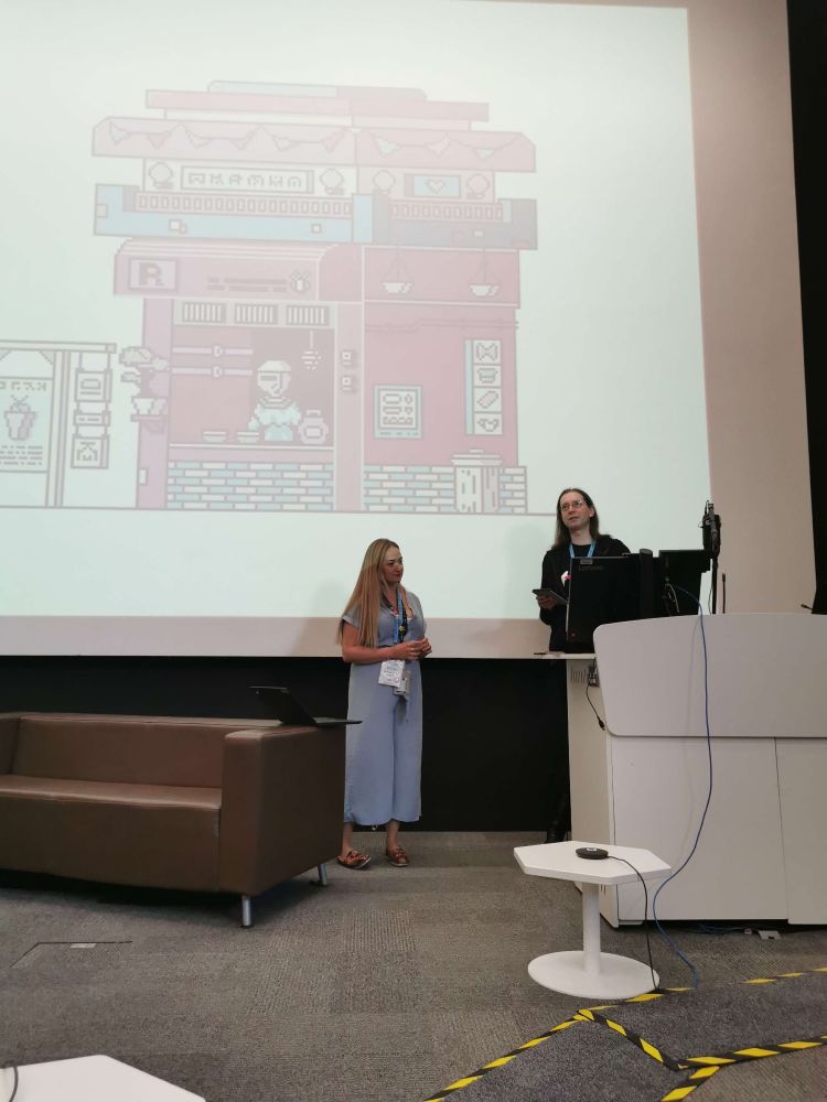 Two people standing at the front of a lecture theatre, next to a brown couch, with a screenshot of a video game projected on the background wall.