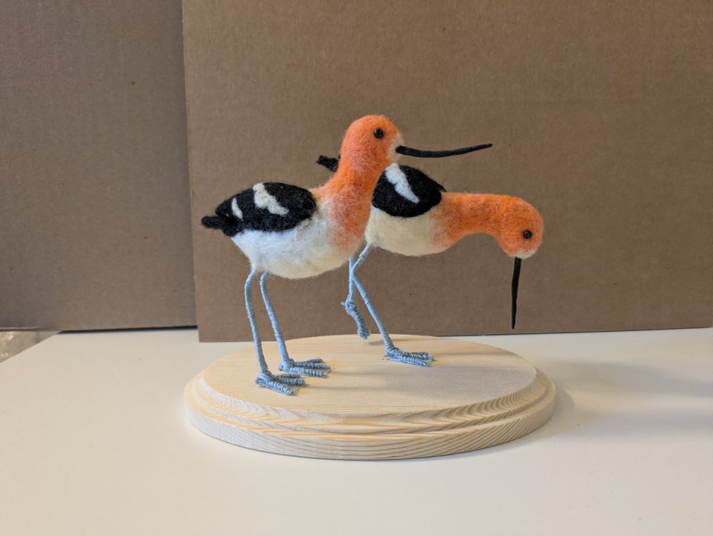 A felt sculpture of two birds with long necks and bills. They have black and white wings and orange heads