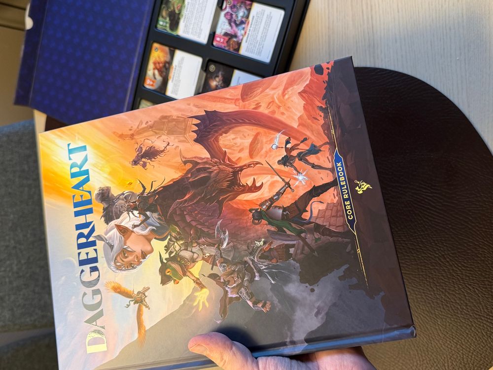 A hand holding a book to show it. The book is the core rulebook for the tabletop roleplaying game daggerheart. Is has a beautiful cover with and elf, a scary monster and other heroic characters. You can see the ability cards in the background.