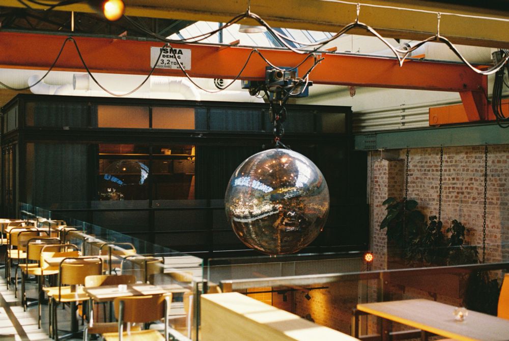 A large disco ball hanging from an industrial crane in a converted factory. There are tables with chairs to the right and sunlight from a skylight above