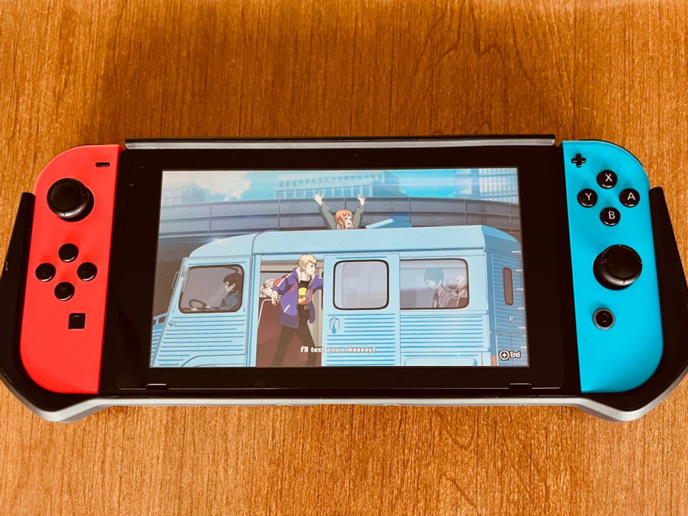 A Nintendo Switch lays on a wood table, on the screen a van drives down the road with teens waving out the windows and doors. Ending cutscene of Persona 5 Royal