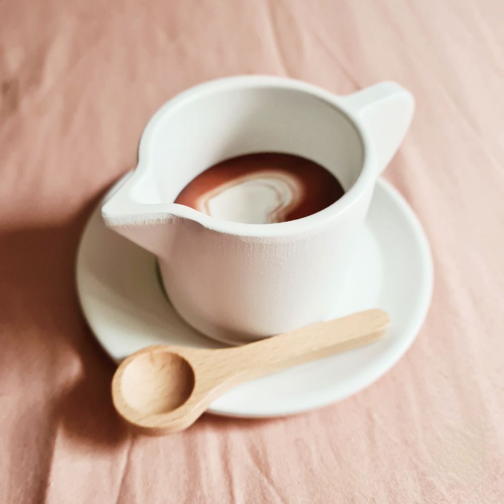 Eine Milchkanne aus Holz auf einer Untertasse auf Holz. In der Milchkanne ist eine runde Holzscheibe, die wie Kaffee aussieht und auf der Untertasse liegt ein kleiner Holzköffel. Das Ganze liegt auf einem lachsfarbenen Bettlaken.
