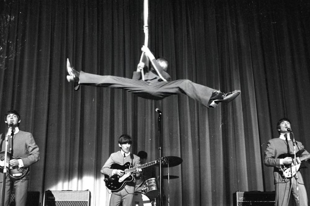 A black and white photograph showing a 1960s rock band performing on stage with a theatrical stunt. A performer is suspended horizontally in mid-air holding onto a stage pole or stand, while other band members in matching suits play guitars and perform below. A drum kit is visible in the background, and the stage has a dark curtain backdrop.